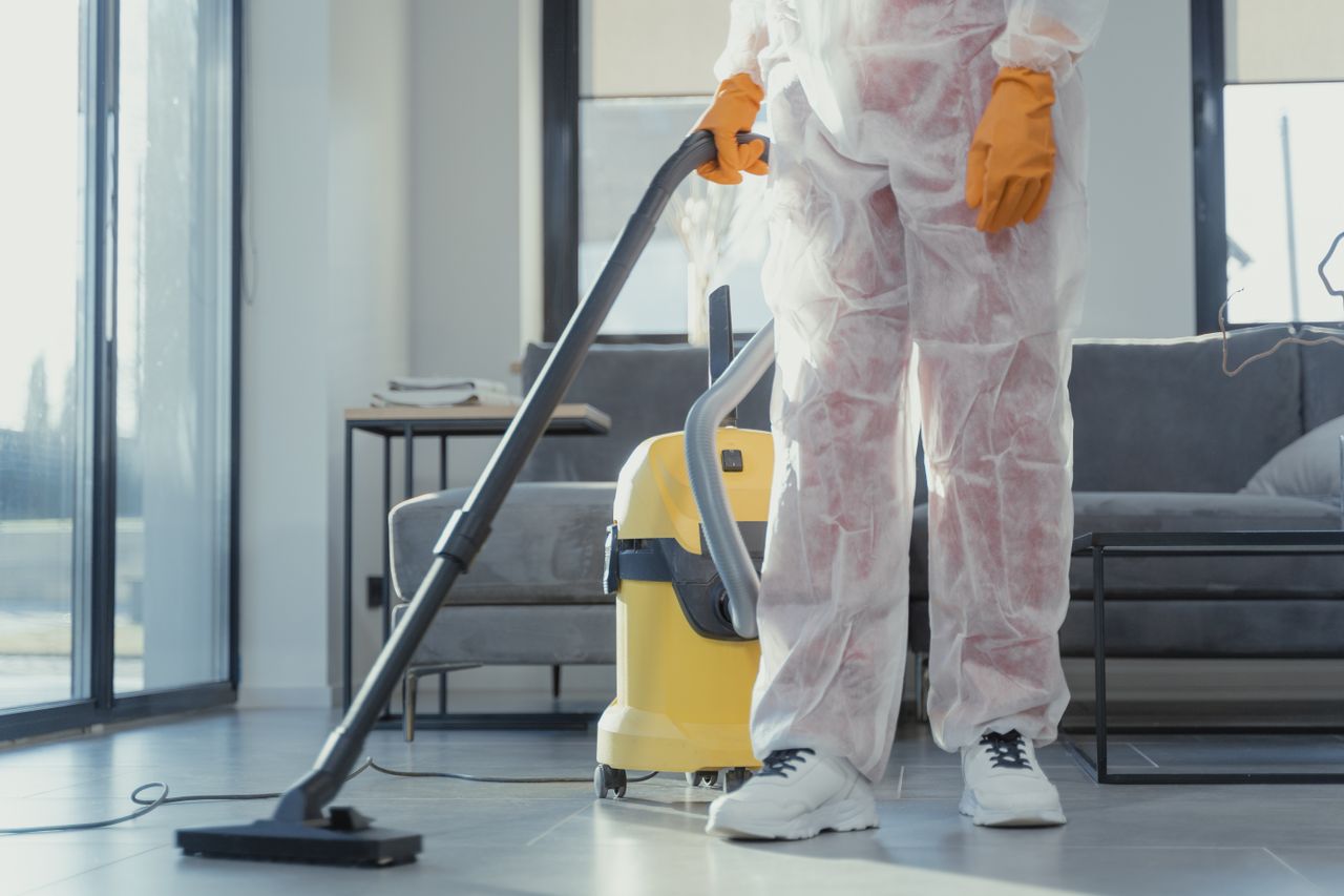 about-img A cleaner in protective coveralls and gloves vacuuming a modern living room.