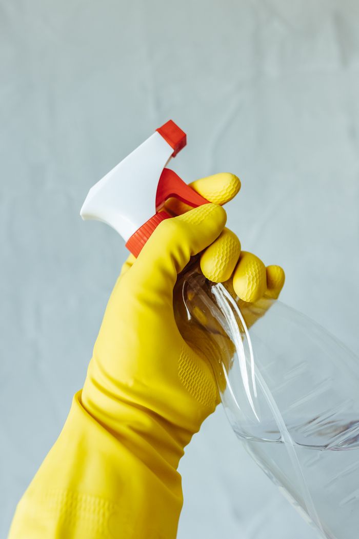 hero-img-01 A hand wearing a yellow rubber glove holds a red and white spray bottle against a neutral background.